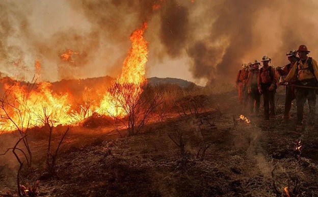 A group of California firefighters on the right, wearing red-and-white hats and fireproof jackets, walk around a brightly burning fire that is burning on the left. There is a lot of smoke in the background.