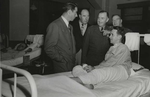 A person in a suit standing next to a person in a hospital bed  AI-generated content may be incorrect.