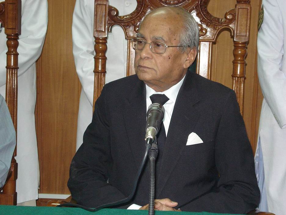 A person in a suit sitting at a microphone Description automatically generated